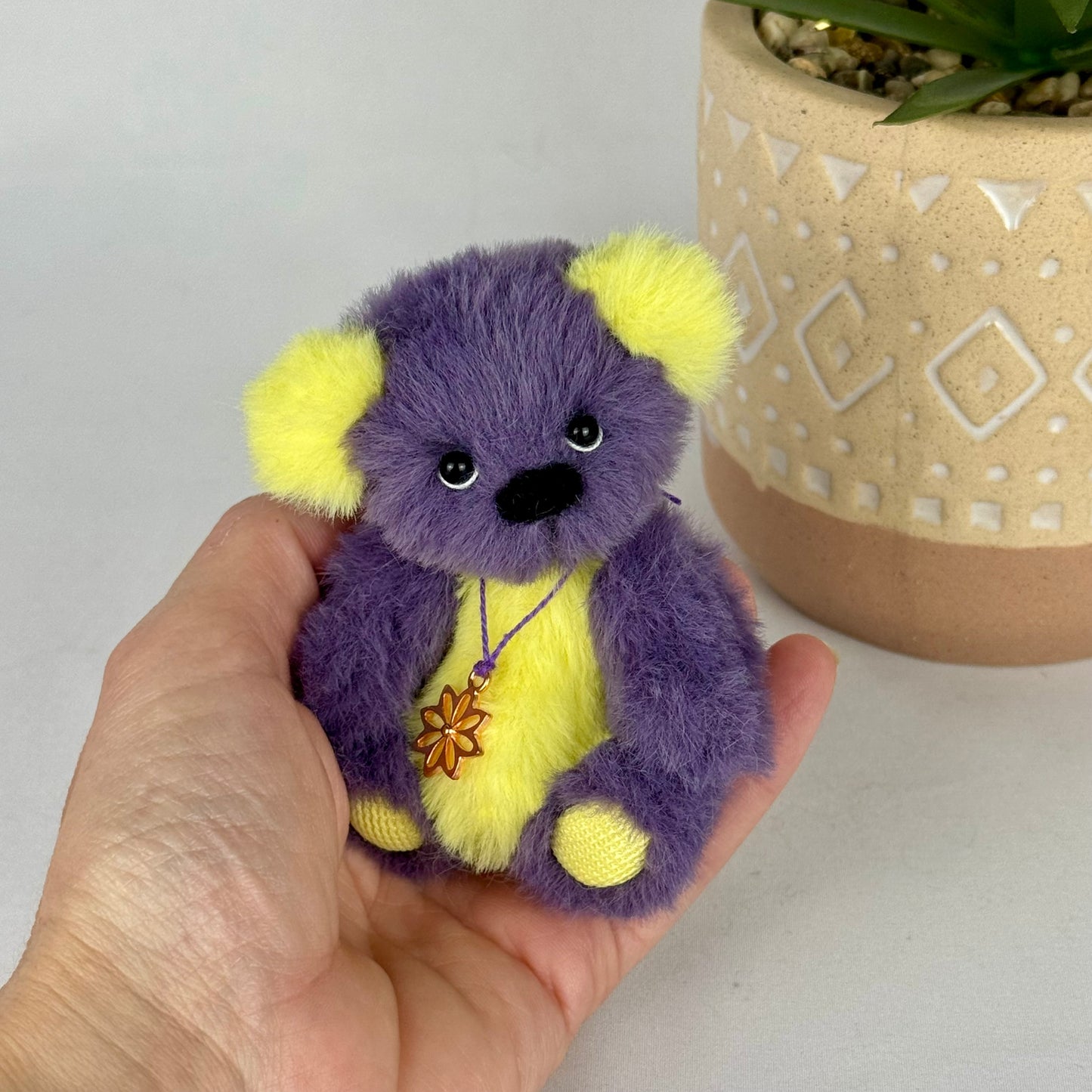 Ours miniature de collection Purple, pelage violet et jaune vif, assis dans une main devant un pot.