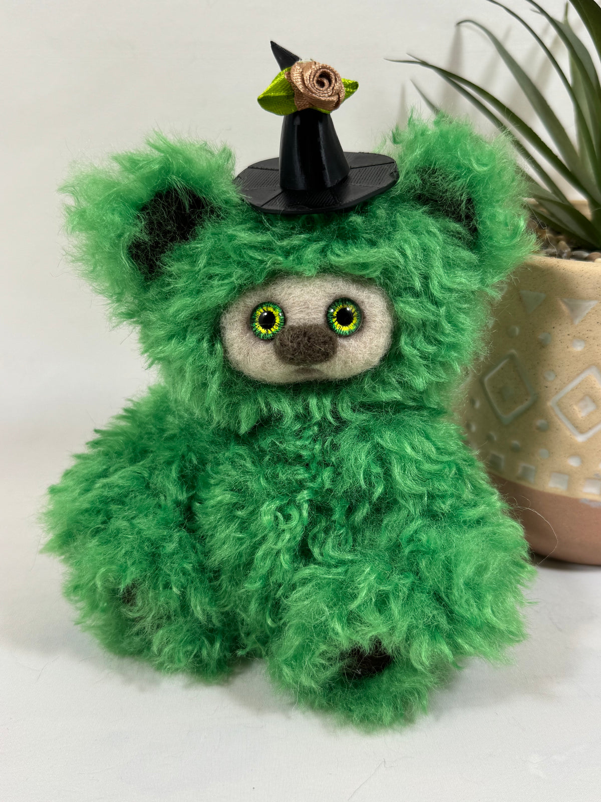 Tambouillette, Frimousse Halloween artisanale de 16 cm en fausse fourrure verte frisée, avec grands yeux verts et jaunes et chapeau de sorcière noir orné d'une rose beige.
