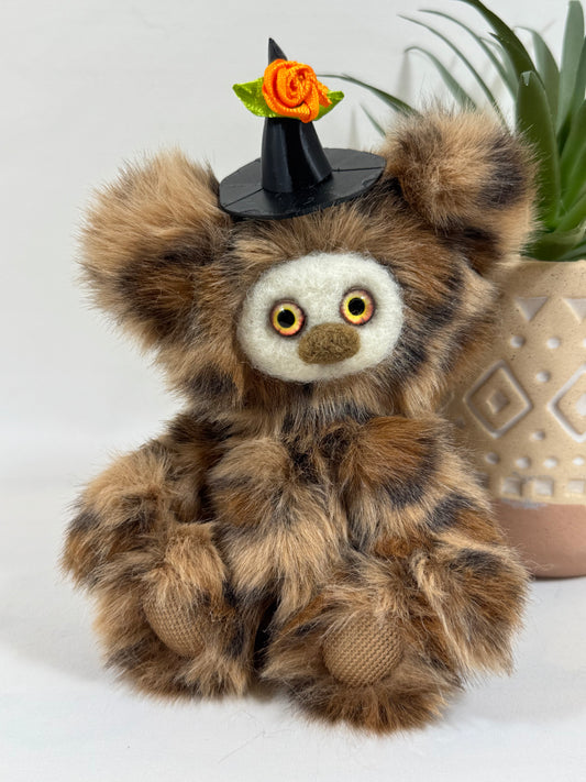 Fripouillette, Frimousse Halloween artisanale de 16 cm en fausse fourrure marron et caramel, avec grands yeux orangés et chapeau de sorcière noir orné d'une rose orange.