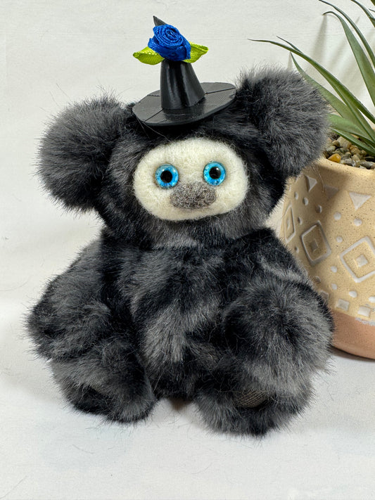 Gribouillette, Frimousse Halloween artisanale de 16 cm en fausse fourrure gris anthracite, avec grands yeux bleu turquoise et chapeau de sorcière noir décoré d'une rose bleue.