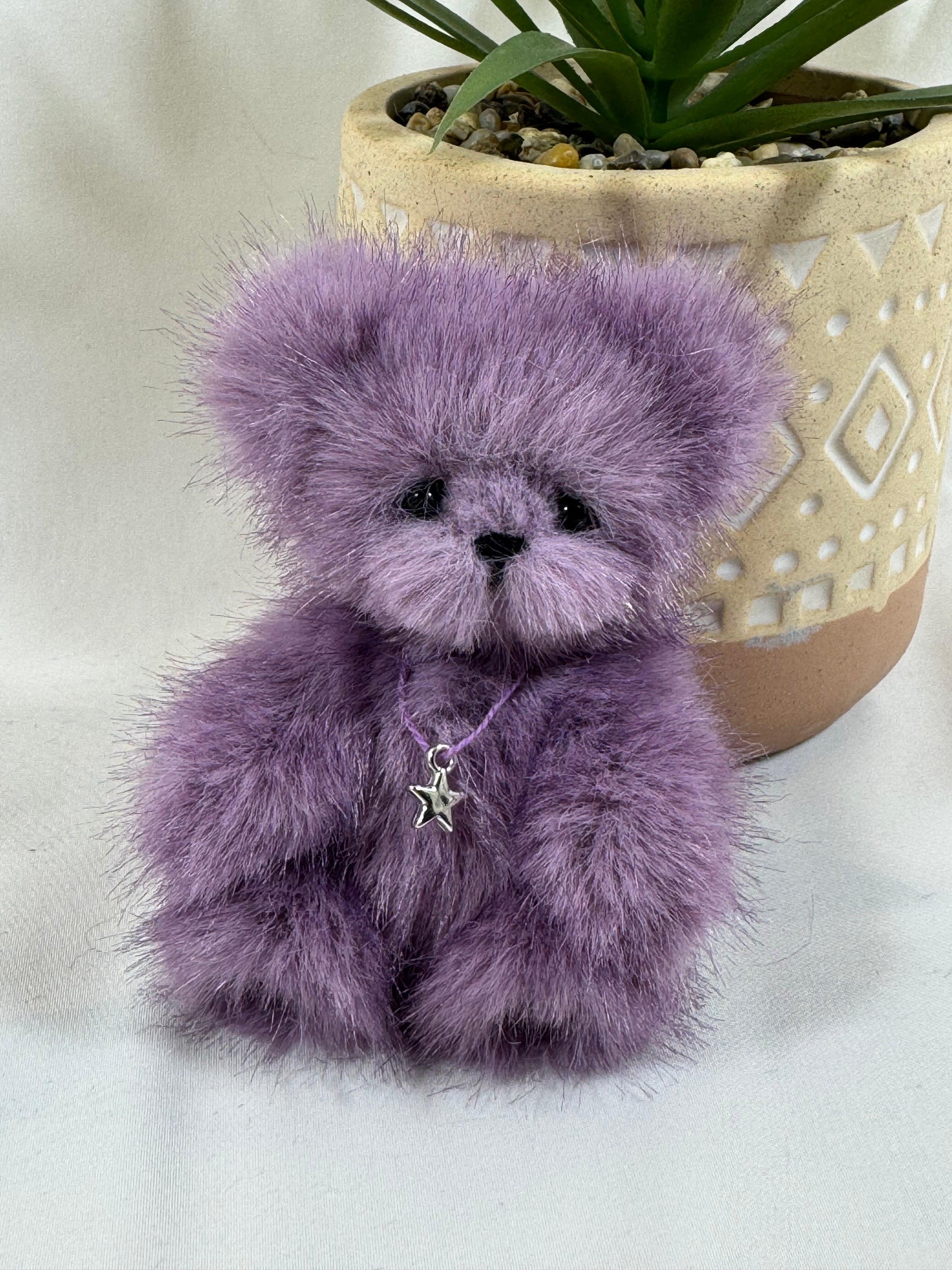 Ours artisanal Stella 10 cm en fausse fourrure violette, près d’une plante.