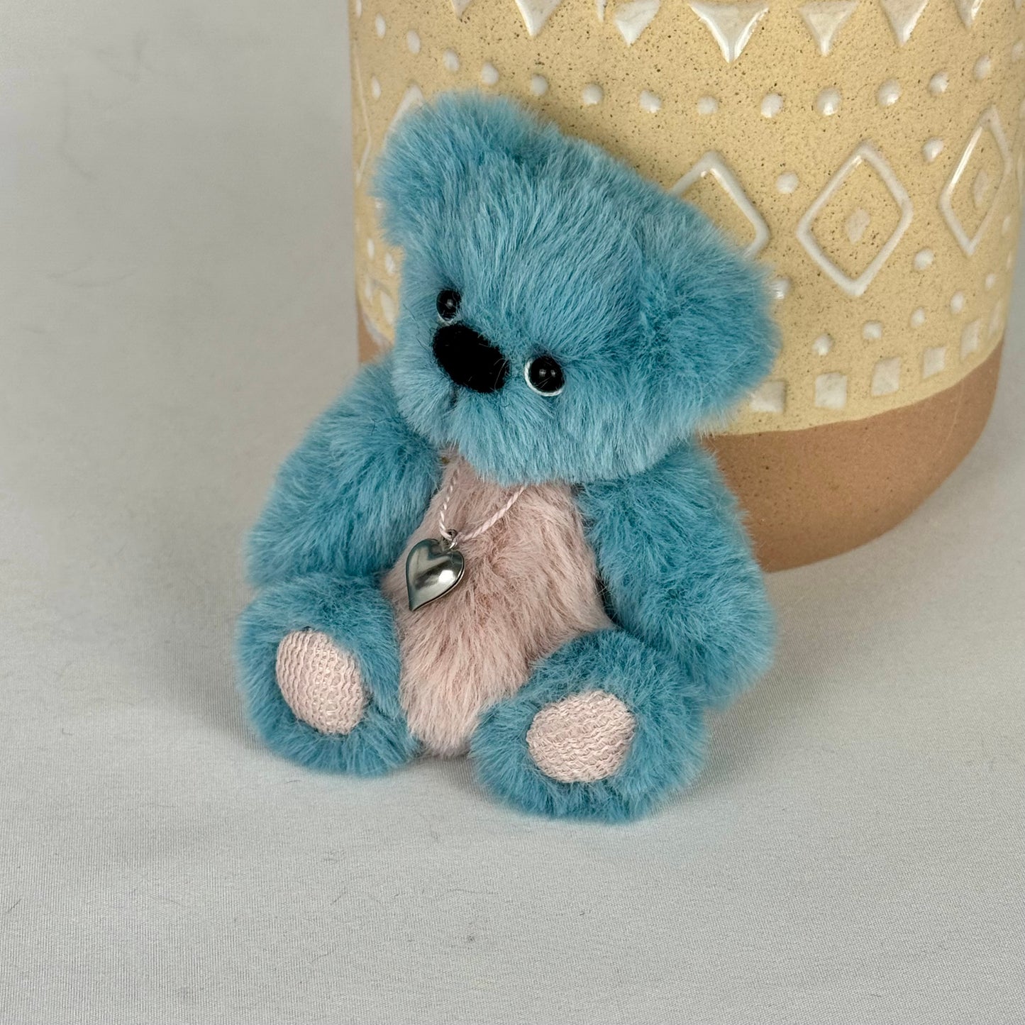 Mini ours de 10 cm, assis devant un pot, yeux en verre et collier cœur en métal.
