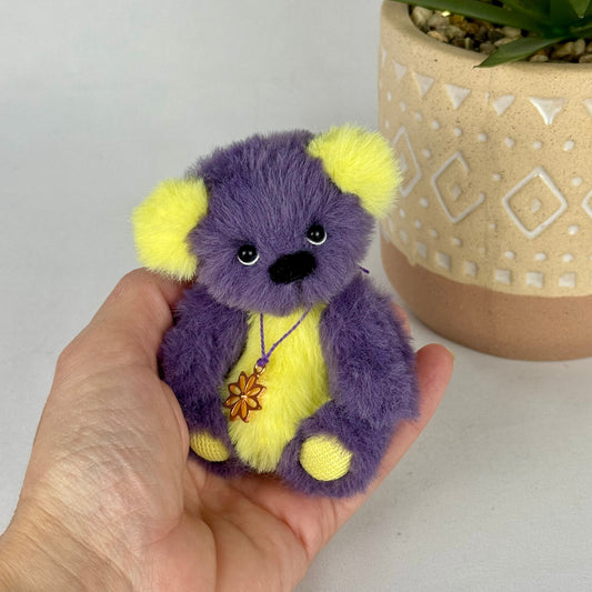 Ours miniature de collection Purple, pelage violet et jaune vif, assis dans une main devant un pot.
